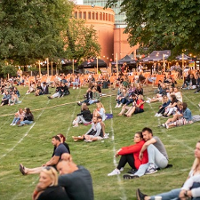 W parku ludzie siedzą na trawie w małych grupkach, najczęściej w parach i najprawdopodobniej słuchają koncertu.