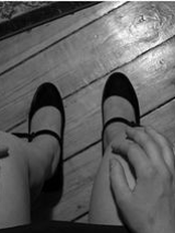 Na czarno-białym zdjęciu będącym kadrem z wystawy widzimy nogi siedzącej w spudnczce kobiety. W tle drewniana podłoga i fragmenty dwóch dywaników.