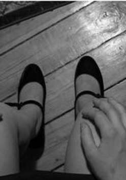 Na czarno-białym zdjęciu będącym kadrem z wystawy widzimy nogi siedzącej w spudnczce kobiety. W tle drewniana podłoga i fragmenty dwóch dywaników.