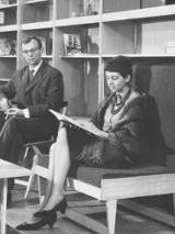 Na czarno-białej fotografii widzimy dwoje ludzi na tle dużego prostego regału. Kobieta znajdująca się bliżej siedzi na drewnianej tapicerowanej ławce będącej częścią regału. Przed nią stoi niewielki, prosty stolik kawowy, a za nią, na krześle, siedzi mężczyzna.