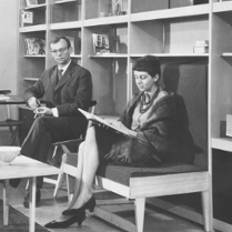 Na czarno-białej fotografii widzimy dwoje ludzi na tle dużego prostego regału. Kobieta znajdująca się bliżej siedzi na drewnianej tapicerowanej ławce będącej częścią regału. Przed nią stoi niewielki, prosty stolik kawowy, a za nią, na krześle, siedzi mężczyzna.