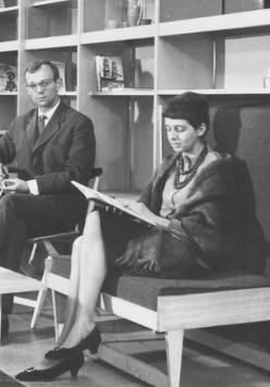 Na czarno-białej fotografii widzimy dwoje ludzi na tle dużego prostego regału. Kobieta znajdująca się bliżej siedzi na drewnianej tapicerowanej ławce będącej częścią regału. Przed nią stoi niewielki, prosty stolik kawowy, a za nią, na krześle, siedzi mężczyzna.