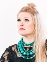 Fotografia przedstawia artystkę. Kobieta ma na sobie czarną suknię. Rękawy są wykonane z koronki, a dół z tiulu. Jasne proste włosy opadają na lewe ramię. Artystka unosi lekko głowę, spogląda poza kadr. Towarzyszą jej instrumenty muzyczne. Postać kobiety wyróżnia się z białego tła.