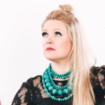 Fotografia przedstawia artystkę. Kobieta ma na sobie czarną suknię. Rękawy są wykonane z koronki, a dół z tiulu. Jasne proste włosy opadają na lewe ramię. Artystka unosi lekko głowę, spogląda poza kadr. Towarzyszą jej instrumenty muzyczne. Postać kobiety wyróżnia się z białego tła.