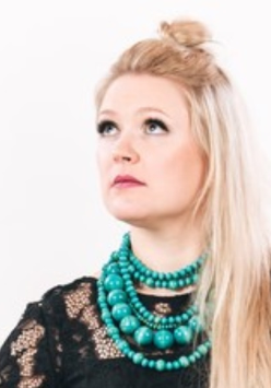 Fotografia przedstawia artystkę. Kobieta ma na sobie czarną suknię. Rękawy są wykonane z koronki, a dół z tiulu. Jasne proste włosy opadają na lewe ramię. Artystka unosi lekko głowę, spogląda poza kadr. Towarzyszą jej instrumenty muzyczne. Postać kobiety wyróżnia się z białego tła.