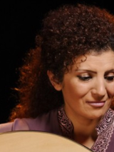 Fotografia przedstawiająca popiersie kobiety ubranej w powłóczystą suknię. Jej spokojną, piękną twarz o regularnych rysach zdobi delikatny uśmiech. Wzrok skupiony ma na trzymanym w rękach owalnym instrumencie strunowym - ud.