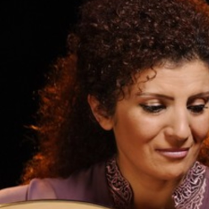 Fotografia przedstawiająca popiersie kobiety ubranej w powłóczystą suknię. Jej spokojną, piękną twarz o regularnych rysach zdobi delikatny uśmiech. Wzrok skupiony ma na trzymanym w rękach owalnym instrumencie strunowym - ud.