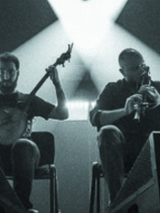 Czarno-białe zdjęcie wykonane zostało podczas koncertu, spod sceny. W centralnej jego części siedzi dwóch muzyków - jeden gra na banjo, drugi na dudach. Po prawej i po lewej ich stronie, nieco w głębi, znajdują się zastawione sprzętem elektronicznym stoły przy których stoi kolejnych dwóch muzyków. W głębi sceny tańczą snopy scenicznych reflektorów.