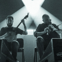 Czarno-białe zdjęcie wykonane zostało podczas koncertu, spod sceny. W centralnej jego części siedzi dwóch muzyków - jeden gra na banjo, drugi na dudach. Po prawej i po lewej ich stronie, nieco w głębi, znajdują się zastawione sprzętem elektronicznym stoły przy których stoi kolejnych dwóch muzyków. W głębi sceny tańczą snopy scenicznych reflektorów.