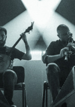 Czarno-białe zdjęcie wykonane zostało podczas koncertu, spod sceny. W centralnej jego części siedzi dwóch muzyków - jeden gra na banjo, drugi na dudach. Po prawej i po lewej ich stronie, nieco w głębi, znajdują się zastawione sprzętem elektronicznym stoły przy których stoi kolejnych dwóch muzyków. W głębi sceny tańczą snopy scenicznych reflektorów.