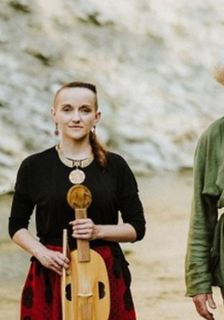 Fotografia ukazuje troje muzyków, dwie kobiety oraz mężczyznę, w tle znajduje się skaliste wzgórze. Cała trójka stoi przed aparatem trzymając instrumenty muzyczne, viola da gamba, vielle oraz lirę korbową. Łagodnie się uśmiechają.