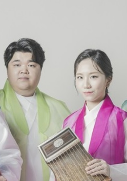 Kolorowa fotografia ukazuje cztery osoby pochodzenia koreańskiego, stojące na wprost aparatu. Trzech mężczyzn oraz kobieta ubrani są w tradycyjne koreańskie stroje. Dwoje muzyków trzyma w dłoniach instrumenty którymi są cytry. Składają się z płaskiego pudła oraz strun.