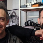 Na pierwszym planie zdjęcia znajduje się dwóch uśmiechniętych mężczyzn Justin Adams & Mauro Durante. Zwróceni są na wprost obiektywu. Na drugim planie znajduje się regał, plakat z dwiema postaciami oraz tarcza o tajemniczych wzorach.