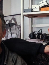 Na pierwszym planie zdjęcia znajduje się dwóch uśmiechniętych mężczyzn Justin Adams & Mauro Durante. Zwróceni są na wprost obiektywu. Na drugim planie znajduje się regał, plakat z dwiema postaciami oraz tarcza o tajemniczych wzorach.