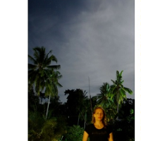 Na zdjęciu widać rezydentkę na tle tropikalnej roślinności i nieba. Widać palmy i wysokie drzewa. Ostrość zdjęcia skierowana jest na tło, a nie na artystkę, która jest na zdjęciu niewyraźna.