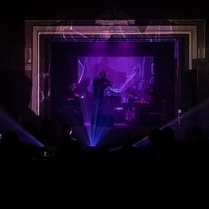 Ciemna fotografia została wykonana na sali koncertowej. W jej centrum widzimy niewyraźnie sylwetkę skrzypka stojącego i grającego na scenie, a za nim sylwetki kilku innych muzyków. Zdjęcie zrobiono ponad głowami osób siedzących na widowni. Utrzymane jest fioletowych barwach światła padającego spod sceny.