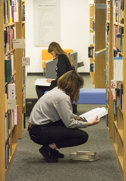 Fotografia dwóch kobiet kucających między regałami z książkami i czytającymi książki.