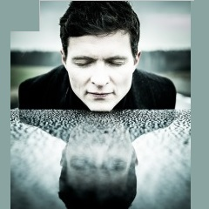 Portret muzyka w zbliżeniu. Jego głowa spoczywa na fortepianie, na powierzchni dostrzec można jej lustrzane odbicie. W tle otwarty leśny krajobraz.