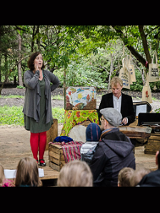 Dwoje aktorów, kobieta i mężczyzna, w trakcie spektaklu. Przed nimi grupa osób oglądających spektakl, w tle park i drzewa.