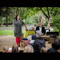 Dwoje aktorów, kobieta i mężczyzna, w trakcie spektaklu. Przed nimi grupa osób oglądających spektakl, w tle park i drzewa.