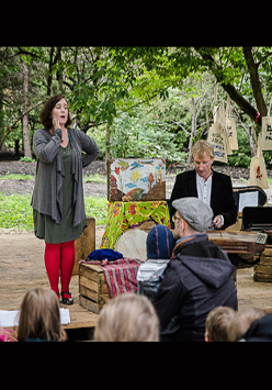 Dwoje aktorów, kobieta i mężczyzna, w trakcie spektaklu. Przed nimi grupa osób oglądających spektakl, w tle park i drzewa.