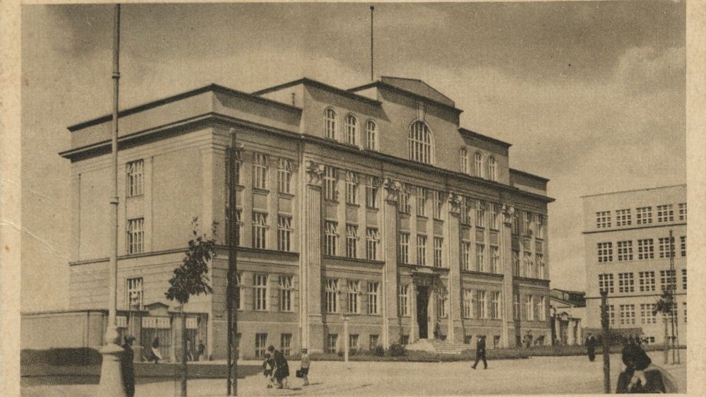 Czarno-białe zdjęcie dużego budynku szkoły w formie pocztówki. Przed budynkiem drzewo i ludzie.