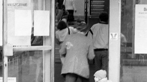 Ludzie wchodzą do punktu wyborczego przez otwarte szklane drzwi. Na pierwszym planie plecy mężczyzny w garniturze, tuż za nim dziecko jadące na zabawkowym rowerku.