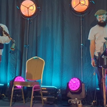 Dwóch mężczyzn na scenie. Jeden gra na dudach a drugi na bębnie. Na scenie stoją też lampy z pomarańczowym i różowym światłem. Z tyłu turkusowa kotara.