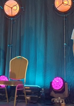 Dwóch mężczyzn na scenie. Jeden gra na dudach a drugi na bębnie. Na scenie stoją też lampy z pomarańczowym i różowym światłem. Z tyłu turkusowa kotara.