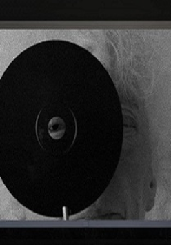 Czarno-biała grafika. Na pierwszym planie widać czarną okrągłą płytę z otworem w samym jej środku. Przez otwór spogląda ludzkie oko. W dalszej perspektywie widać, że za płytą stoi rezydentka.