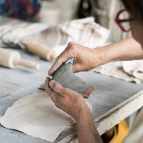 Kobieta modelująca ręcznie gliniany przedmiot na stole warsztatowym, otoczona narzędziami do ceramiki i materiałami do pracy.