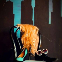 Na czarnym krześle stoi czarny męski but, na nim leżą różowe okulary, obok stoi damki zielony but na wysokim obcasie. Na oparciu krzesła wisi peruka z długimi blond włosami.