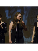 Zdjęcie przedstawia trzy kobiety, w czarnych sukienkach, stojące na scenie podczas koncertu. Przed nimi stoją mikrofony.