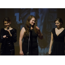 Zdjęcie przedstawia trzy kobiety, w czarnych sukienkach, stojące na scenie podczas koncertu. Przed nimi stoją mikrofony.