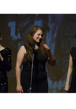 Zdjęcie przedstawia trzy kobiety, w czarnych sukienkach, stojące na scenie podczas koncertu. Przed nimi stoją mikrofony.