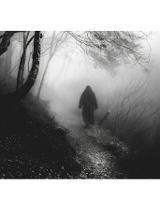 Czarno-białe zdjęcie. Ścieżką w lesie idzie postać ubrana w szeroką czarną szatę z kapturem. Wokół drzewa i krzewy we mgle.