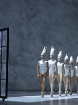 na zdjęciu po lewej stronie chłopiec patrzący przez duże okno, przed oknem rząd baletnic w białych strojach typu body i białych nakryciach głowy