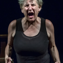 na zdjęciu kobieta w średnim wieku patrzy w obiekty i krzyczy głośno, ręce ma skierowane do przodu, place zagięte