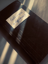 na zdjeciu widoczny czarny zeszyt, na okładce w gornej części naklejony prostokąt z kartki zeszytowej w kratkę, z napisem "Ćwiczenia z dyplomacji"