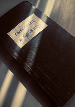 na zdjeciu widoczny czarny zeszyt, na okładce w gornej części naklejony prostokąt z kartki zeszytowej w kratkę, z napisem "Ćwiczenia z dyplomacji"