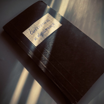 na zdjeciu widoczny czarny zeszyt, na okładce w gornej części naklejony prostokąt z kartki zeszytowej w kratkę, z napisem "Ćwiczenia z dyplomacji"