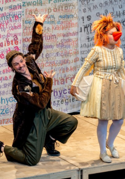 po lewej stronie zdjęcia klęczący na jedno kolano mężczyzna w czapce, ubrany w długi futrzany brązowy płaszcz, po prawej stojąca kobieta w sukience, rudej peruce i pomarańczowym doklejonym dziobie