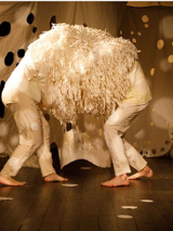 grafika przedstawia dwie bose, pochylone postaci stojące przodem do siebie, na głowach i barkach mają narzuconą narzutę z długimi frędzlami