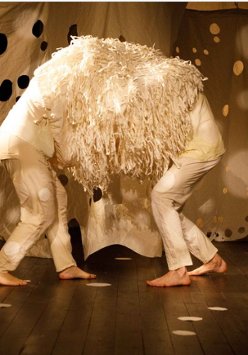 grafika przedstawia dwie bose, pochylone postaci stojące przodem do siebie, na głowach i barkach mają narzuconą narzutę z długimi frędzlami