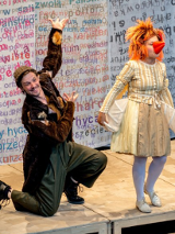 po lewej stronie zdjęcia klęczący na jedno kolano mężczyzna w czapce, ubrany w długi futrzany brązowy płaszcz, po prawej stojąca kobieta w sukience, rudej peruce i pomarańczowym doklejonym dziobie