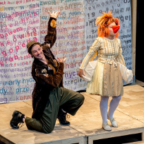 po lewej stronie zdjęcia klęczący na jedno kolano mężczyzna w czapce, ubrany w długi futrzany brązowy płaszcz, po prawej stojąca kobieta w sukience, rudej peruce i pomarańczowym doklejonym dziobie