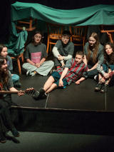 na zdjęciu grua młodych osób siedzi na podłodze, jedna z nich gra na gitarze, za nimi stoją krzesła z narzuconym na oparcia kocem