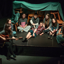 na zdjęciu grua młodych osób siedzi na podłodze, jedna z nich gra na gitarze, za nimi stoją krzesła z narzuconym na oparcia kocem