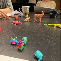 na zdjęciu widoczny stół z leżącymi kolorowymi figurkami zwierząt, za stołem widoczne fragmenty dwóch siedzących postaci