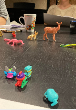 na zdjęciu widoczny stół z leżącymi kolorowymi figurkami zwierząt, za stołem widoczne fragmenty dwóch siedzących postaci